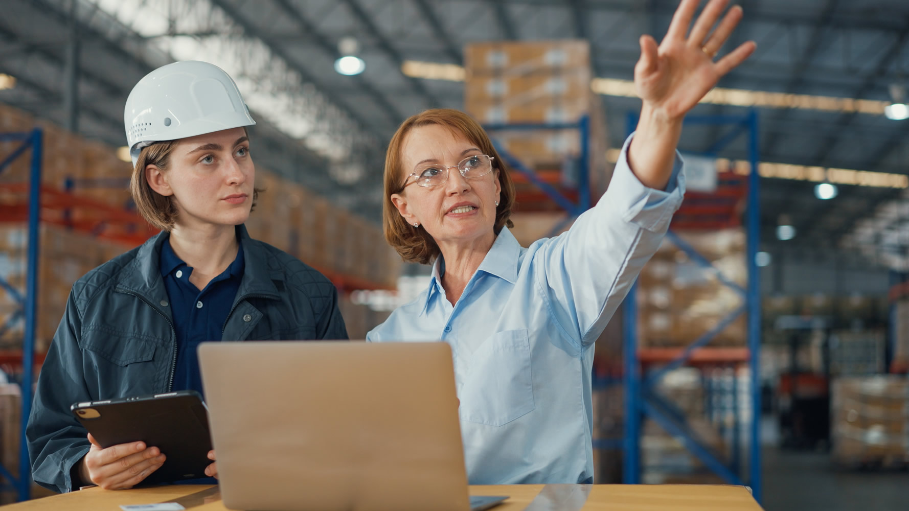 Young caucasian woman worker and mature female manager checks stock inventory with tablet and laptop and discuss talk together in retail warehouse. Logistic industry business concept.